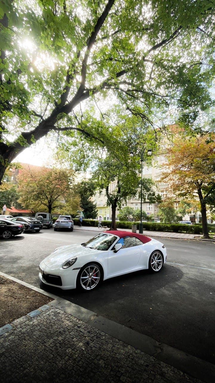 White Porsche 911 Cabriolet with a red soft top parked under green trees on a city street